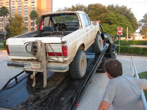1989 Ford Bronco - A white 1989 Ford Bronco is loaded onto a flatbed tow truck, showing its rear end with a mounted spare tire.