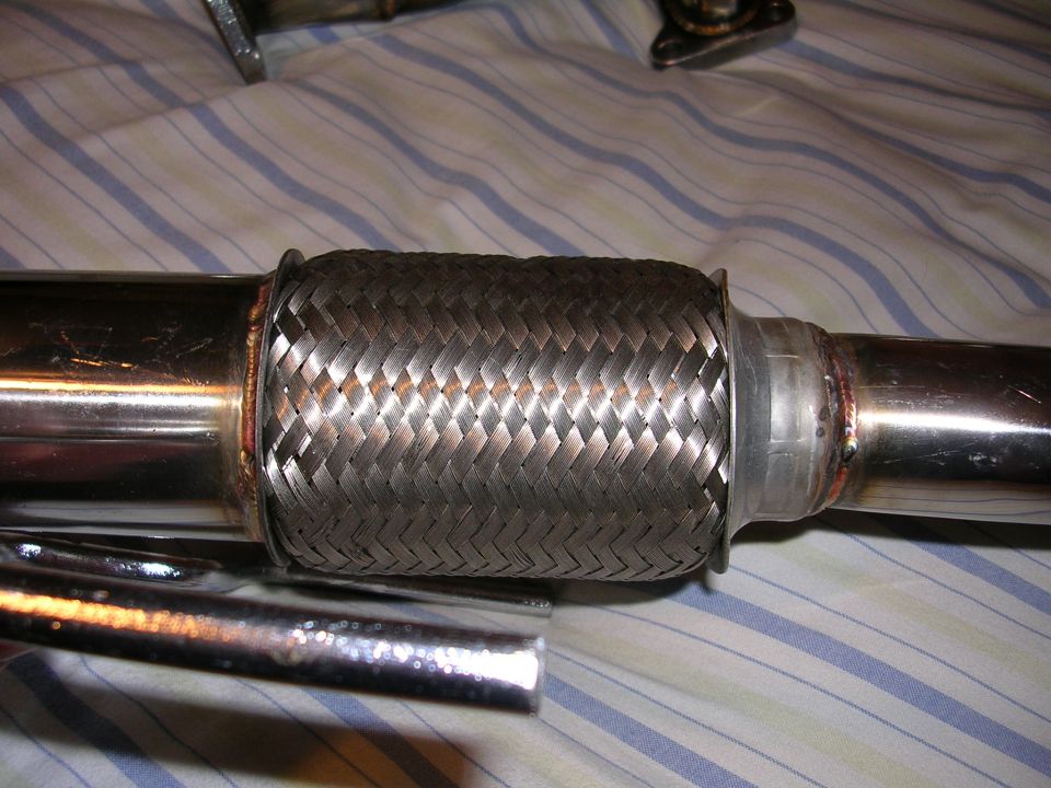 Close-up of a braided metal exhaust flex pipe with shiny metal tubing on either side. The pipe is resting on a striped fabric surface