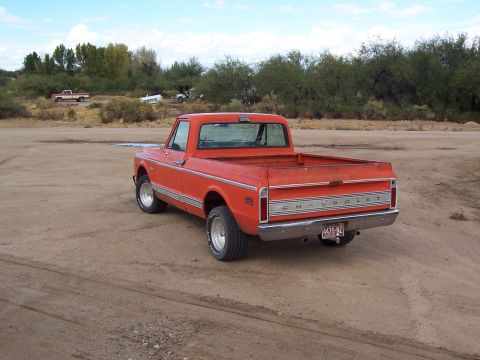 1972 Chevrolet C/K Pickup - An orange pickup truck is parked in a dirt lot with trees and another truck in the background.