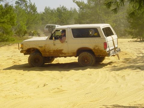 1989 Ford Bronco - Two Ford Broncos navigate a dusty, off-road trail with sandy terrain and scattered trees.