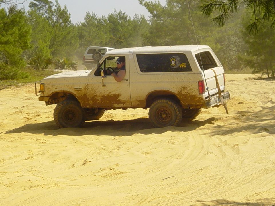 White Ford Bronco driving on a sandy trail, covered in mud. Another Bronco is behind it