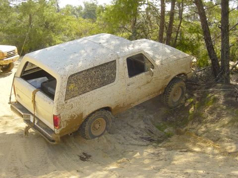 1989 Ford Bronco - A white SUV is covered in mud while navigating a dirt path with trees in the background.