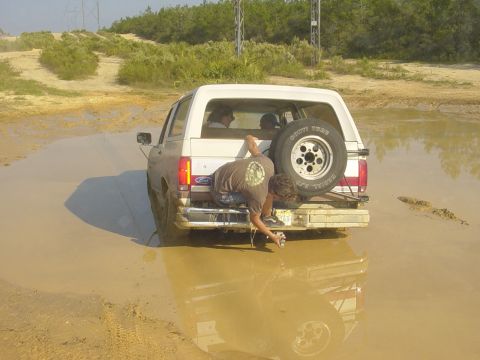 1989 Ford Bronco - A white Ford Bronco is stuck in a muddy puddle with a person attempting to retrieve something from the tailgate.