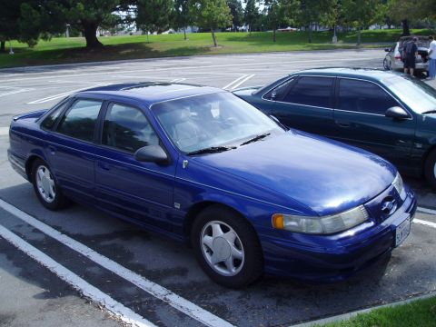 1993 Ford Taurus - A blue sedan is parked in a lot next to another car, with trees and grass in the background.