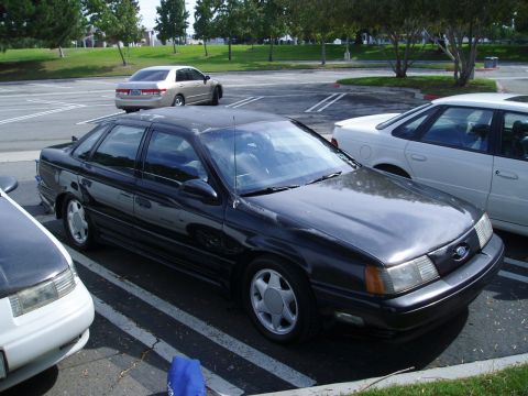 1993 Ford Taurus - A dark sedan is parked in a lot next to other vehicles.