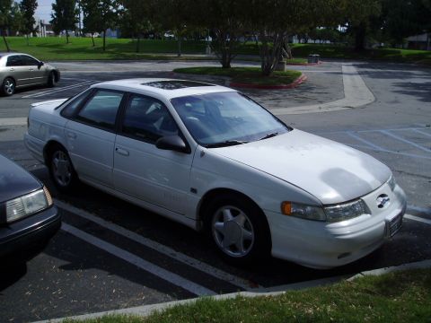 1993 Ford Taurus - A white 1993 Ford Taurus sedan is parked in a lot, with another car visible to the left.