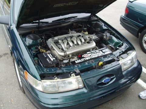 1993 Ford Taurus - Engine bay of a 1993 Ford Taurus SHO, showing the V6 engine and aftermarket parts. A second Taurus is visible in the background.