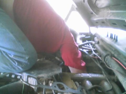 1990 Ford Bronco - person in a red shirt and jeans leans deep into a vehicle's engine bay, working on a tangle of wiring under the open hood.