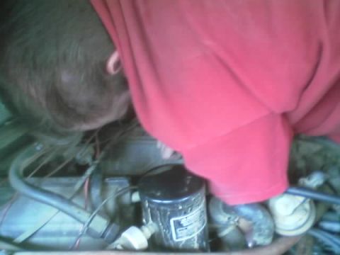 1990 Ford Bronco - person in a red shirt leans head-first into an engine compartment, working near a black oil filter amid hoses and wiring.