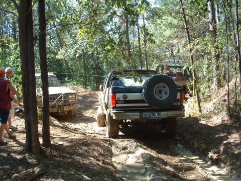 1993 Ford Bronco - Two off-road vehicles navigate a muddy trail in a wooded area.