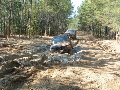 1993 Ford Bronco - Two SUVs, including a 1993 Ford Bronco, struggle through a muddy off-road trail surrounded by pine trees.
