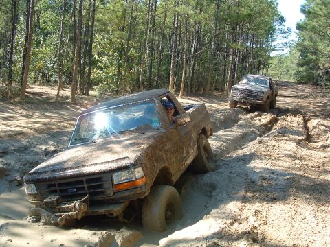 1993 Ford Bronco - Two trucks, a Ford Bronco and a pickup, struggle through a muddy off-road trail in a wooded area.
