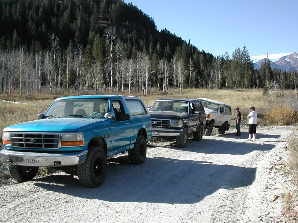 Three Ford Broncos parked on a dirt road with a wooded hillside in the background. The lead Bronco is teal