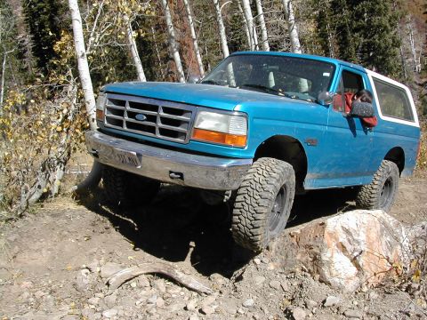 1994 Ford Bronco - A blue Ford Bronco navigates a challenging rocky terrain with trees in the background.