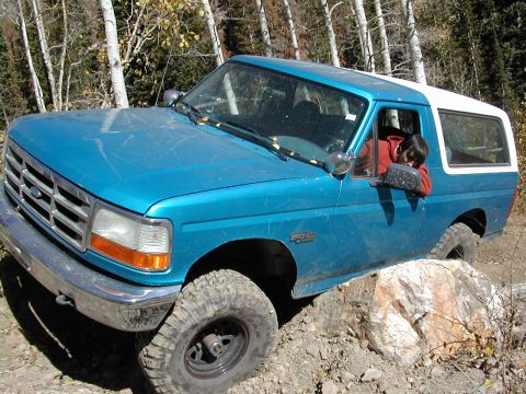 1994 Ford Bronco - A blue 1994 Ford Bronco is shown off-roading on a rocky trail with a person inside.