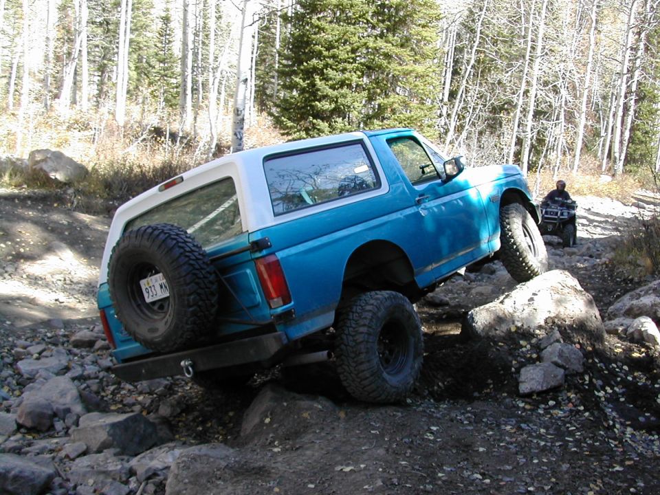 Blue 1994 Ford Bronco climbing over rocks on a dirt trail; ATV visible in background