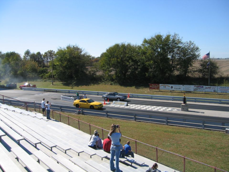 Yellow Mustang and gray convertible staged at a drag strip seen from the stands