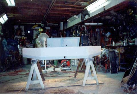1989 Ford Bronco - A wide, smooth front bumper shell finished in white primer sits face-on across two white sawhorses in a cluttered home garage. Behind it ar…