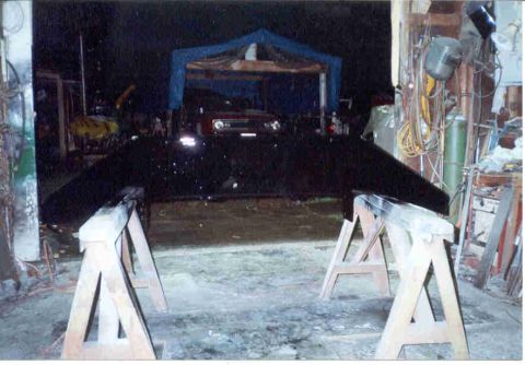 1989 Ford Bronco - A freshly painted black fabricated bumper resting across two wooden sawhorses in a dim shop, its wing-shaped ends angling back toward the f…