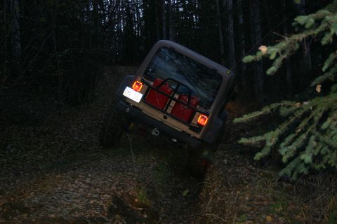 2003 Jeep TJ - A tan Jeep Wrangler drives off-road through a wooded area at dusk, with two jerrycans strapped to its rear.