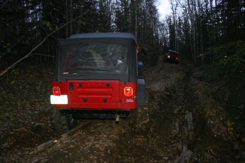 2003 Jeep TJ - A red Jeep Wrangler navigates a muddy, wooded trail, with another vehicle visible further up the path.