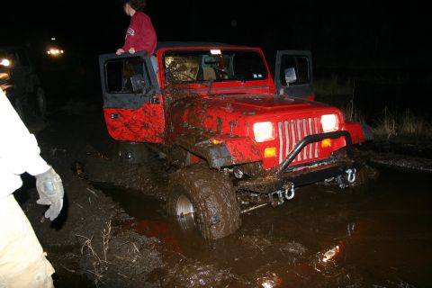 2003 Jeep TJ - A red Jeep TJ is stuck deep in mud with its doors open, while another vehicle is visible in the background.
