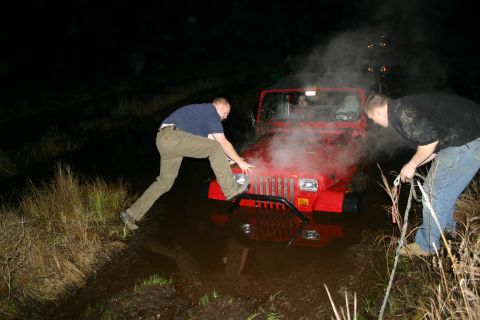2003 Jeep TJ - A red Jeep Wrangler is stuck in muddy water at night, with two men trying to help it.