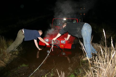2003 Jeep TJ - Two individuals attempt to free a red 2003 Jeep TJ from a muddy ditch at night, with steam rising from the engine.