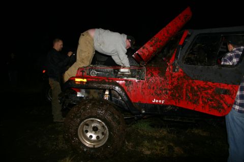 2003 Jeep TJ - Muddy red Jeep with hood open, people attending to the engine at night.