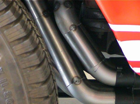 1979 Ford Bronco - Close-up view of exhaust headers and a tire, with a portion of an orange and white striped vehicle body visible.