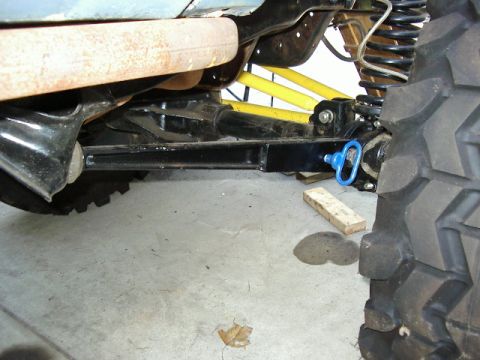 1985 Ford Bronco - The underside of a vehicle shows its suspension, axle, and exhaust system, with a blue tow hook attached.