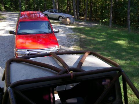 1985 Ford Bronco - A red SUV and a silver coupe are parked on a gravel driveway surrounded by trees.