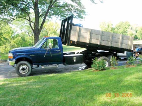 Side view of a blue Ford F-Series dump truck with a wooden bed. The truck has a single cab and dual rear wheels