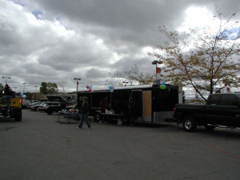 1977 Ford F-150 - A black pickup truck is parked next to a black trailer, with several people milling around in a parking lot.