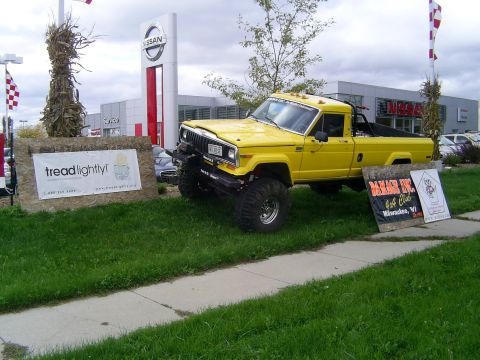 1977 Ford F-150 - Lifted Jeep pickup truck with off-road tires parked near a dealership, with club and environmental group signage.