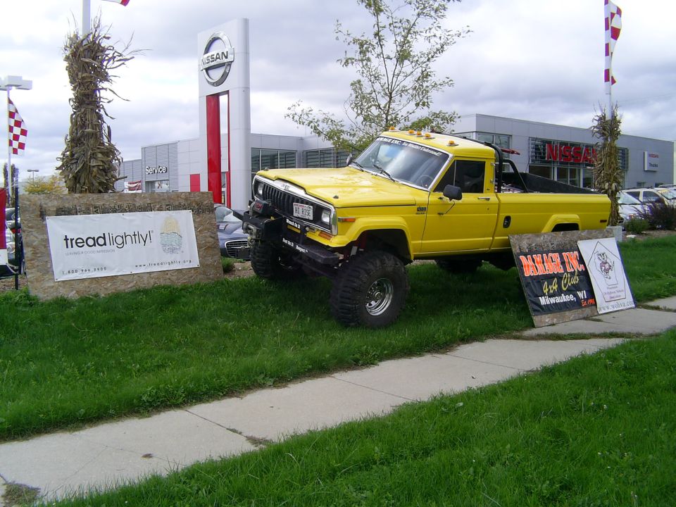 Yellow lifted Jeep pickup truck parked on grass in front of a Nissan dealership. Signs for Tread Lightly and a 4x4 club are visible