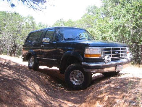 1995 Ford Bronco - This 1995 Ford Bronco is captured off-road, navigating a dusty trail surrounded by trees.