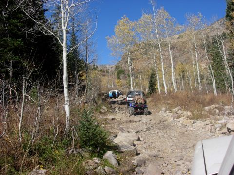 1987 Ford Bronco - Several off-road vehicles navigate a challenging rocky trail through a wooded mountainside during autumn.