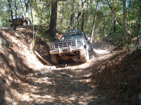 1993 Ford Bronco - A black off-road truck, likely from the mid-1970s, tackles a challenging dirt path in a wooded area, with another vehicle visible on the hi…
