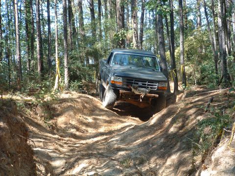 1993 Ford Bronco - A dark pickup truck navigates a challenging off-road trail through a wooded area.
