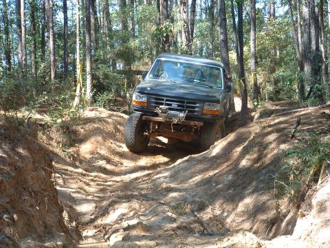 1993 Ford Bronco - A dark SUV navigates a challenging muddy path through a dense forest.