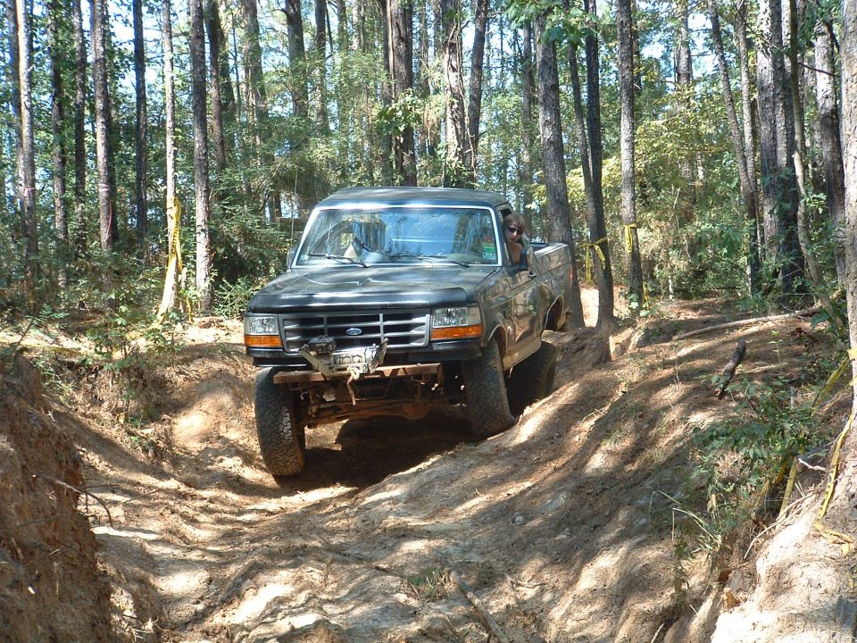 Dark green topless lifted Ford Bronco with winch descending a rutted dirt trail in pine woods