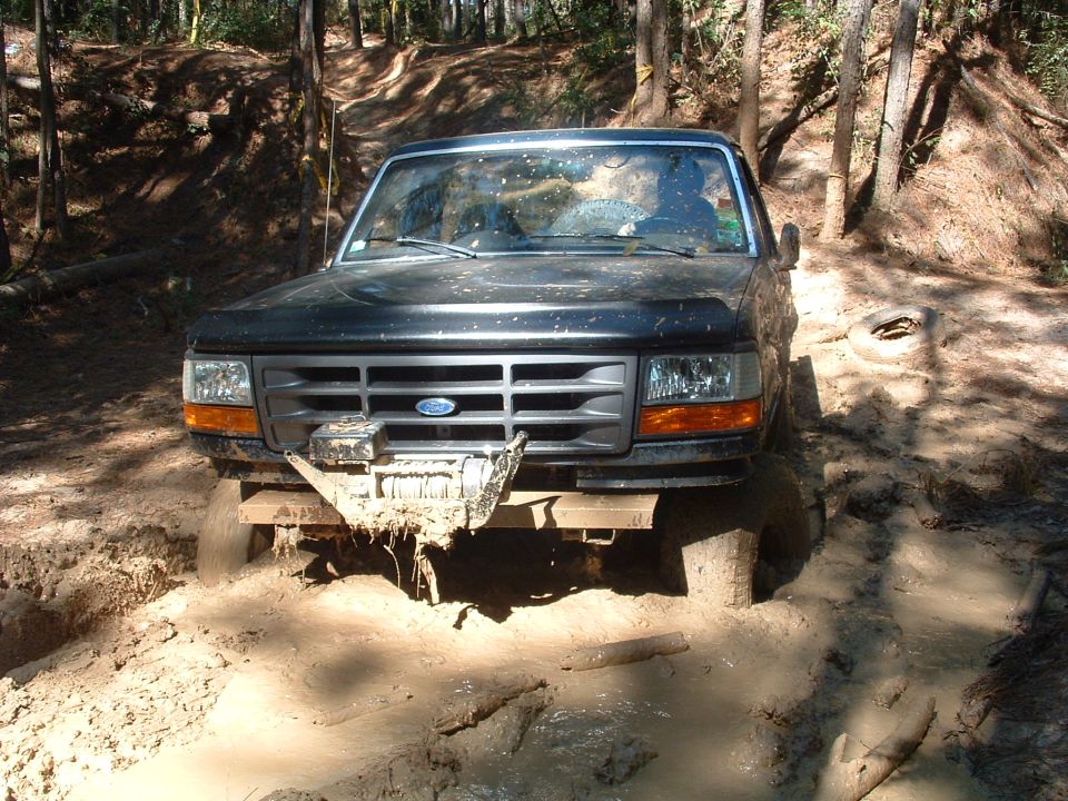 Black 1990s Ford 4x4 buried to the axles in a muddy forest pit, front view