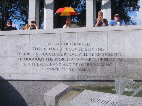 1988 Ford Bronco - A group of people observe a memorial, with an inscription on the stone wall and a colorful umbrella providing shade.