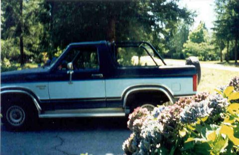 1989 Ford Bronco - Side profile of a two-tone dark blue and white 1980s Ford Bronco with the removable hardtop off, showing an exposed roll bar behind the cab…