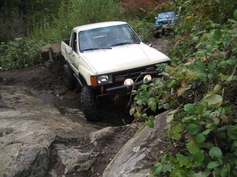 1990 Ford Bronco - Two Toyota trucks off-roading on a challenging, rocky trail with dense foliage.