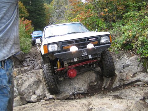 1990 Ford Bronco - A white Toyota pickup truck with off-road modifications is seen navigating a challenging rocky trail, with another blue vehicle following b…
