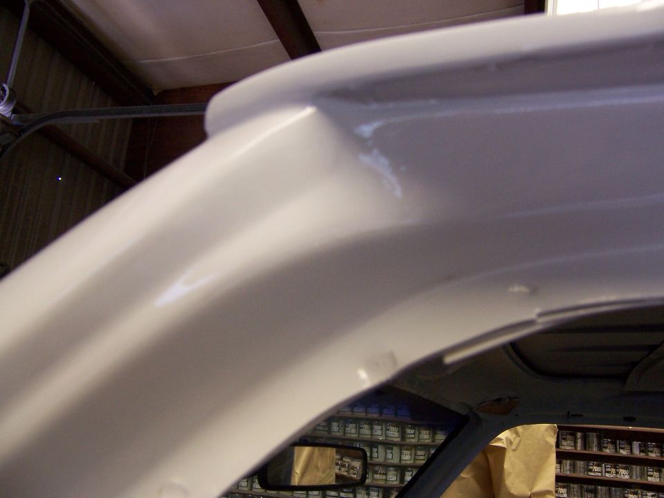Close-up of a vehicle's interior roofline and window frame, showing primer and paint work. Shelves of paint cans are visible in the background