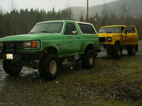 1990 Ford Bronco - Two lifted 4x4s, a green Bronco and a yellow F-Series, sit parked on a gravel lot during a rainy day.