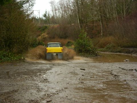 1990 Ford Bronco - A yellow pickup truck navigates a muddy trail, kicking up water and dirt.